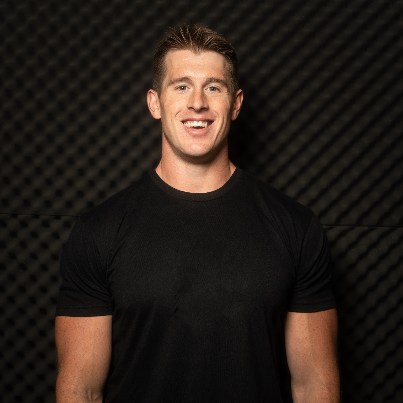 A young man smiling and wearing a black athletic shirt, standing in front of soundproofing foam panels.