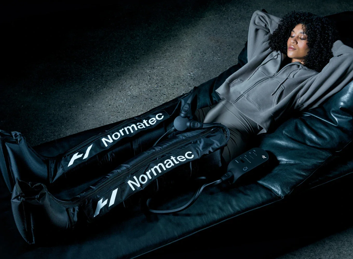 Woman lying on a couch with eyes closed, wearing a gray hoodie, with a motorized leg brace labeled 'Normatec' on her legs, connected to a control device.