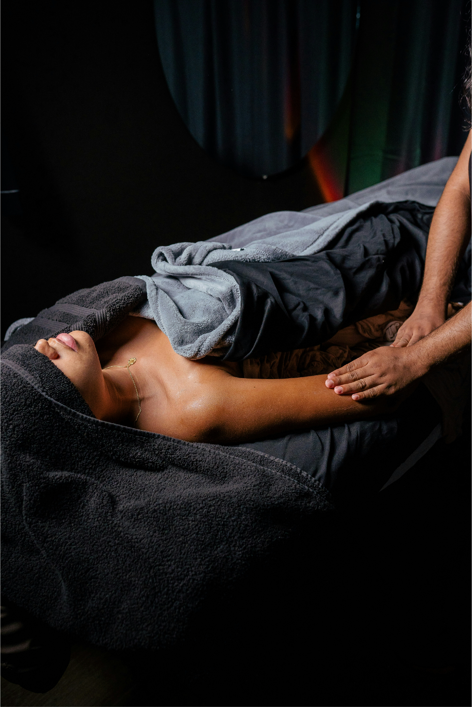 Person receiving a massage, lying on a massage table, with a towel covering their lower body, and another towel draped over their chest, with a massage therapist providing the treatment.
