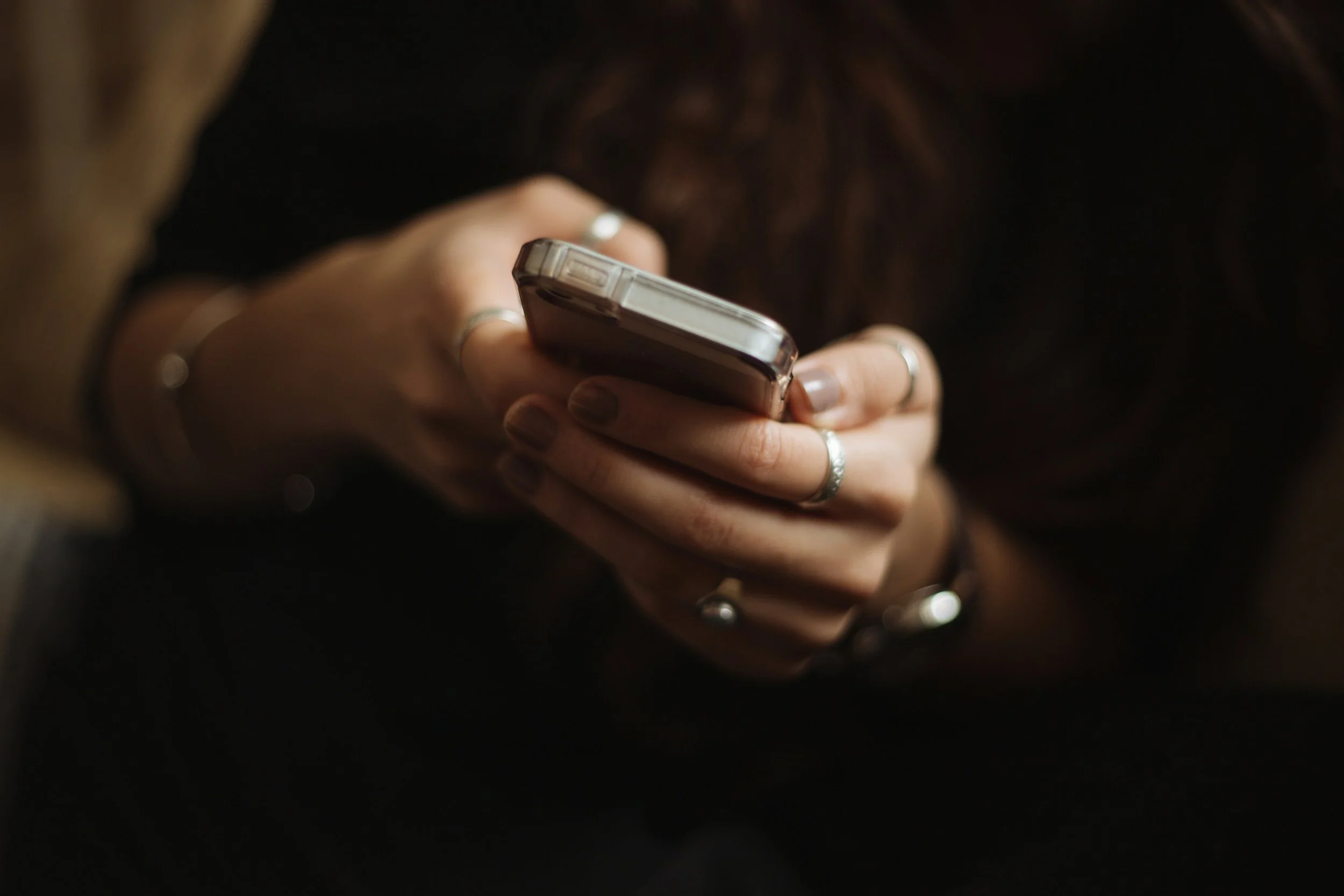 Person holding a smartphone, wearing rings and bracelets, with long brown hair visible.