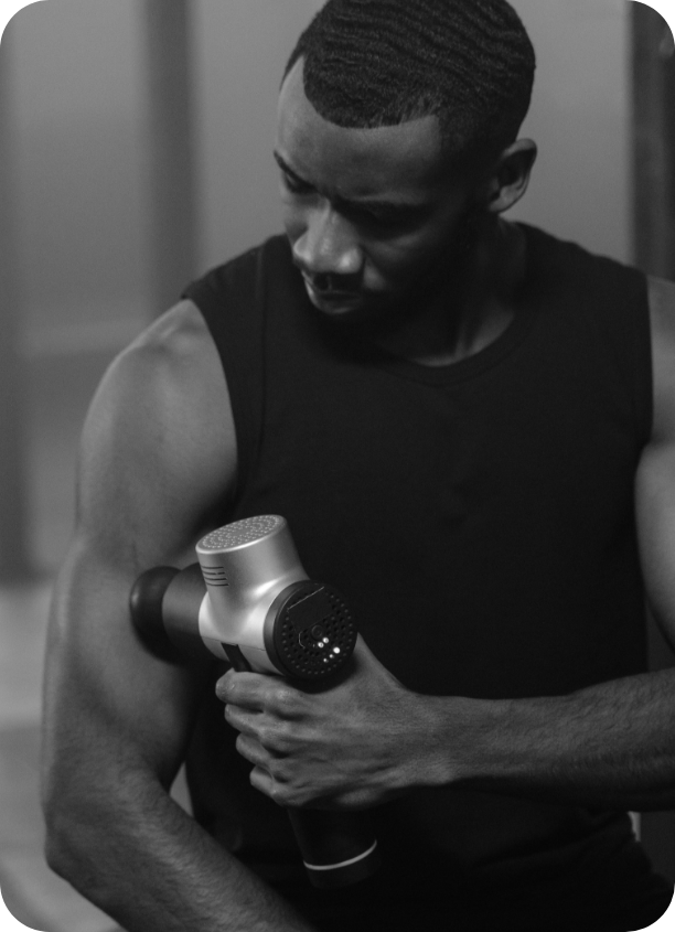 A man in a sleeveless shirt is holding a massage gun against his upper arm, looking down at it in a black-and-white photo.