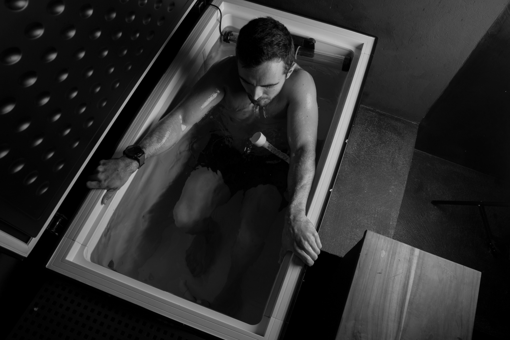 A man sitting in an ice bath with his arms resting on the edge, wearing a smartwatch, and looking down.