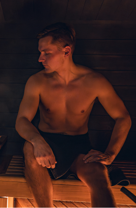 A shirtless man sitting inside a wooden sauna, looking down with a relaxed expression.