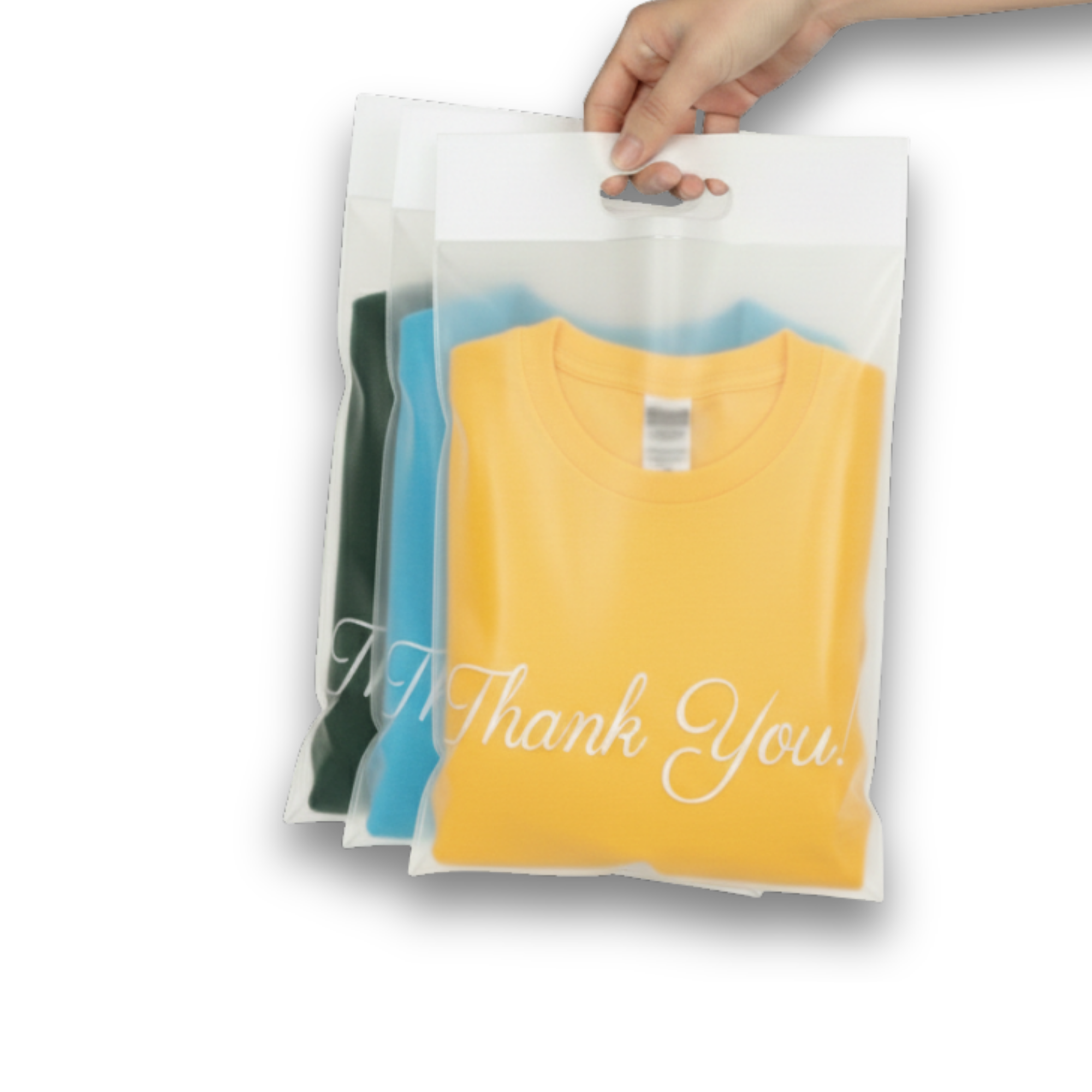 Hand holding three transparent plastic bags with folded shirts inside, one yellow with 'Thank You!' written on it, one blue, and one green.