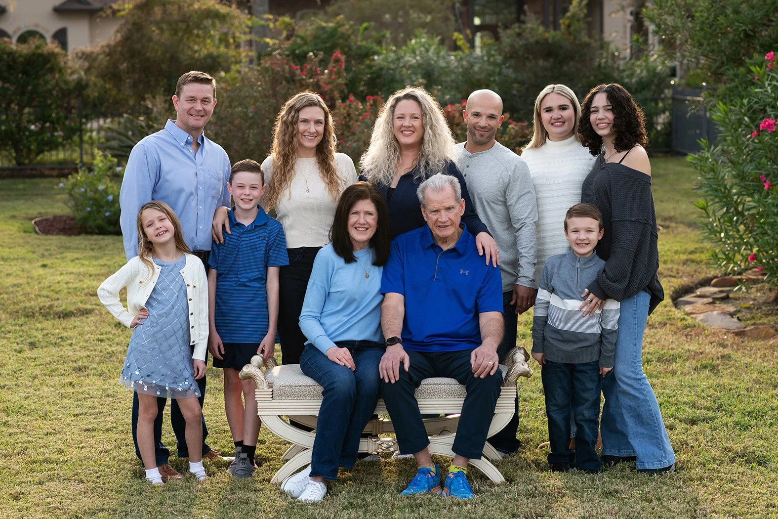 A large multi-generational family of fourteen people gathered outdoors in a garden, posed for a group photo with a mix of adults and children smiling at the camera.