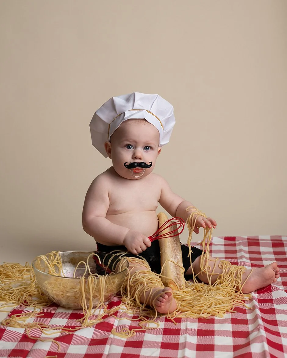 Baby dressed as a chef, with a chef hat and fake mustache, surrounded by spaghetti noodles, holding a whisk, with a bowl of spaghetti, sitting on a checkered red and white tablecloth.