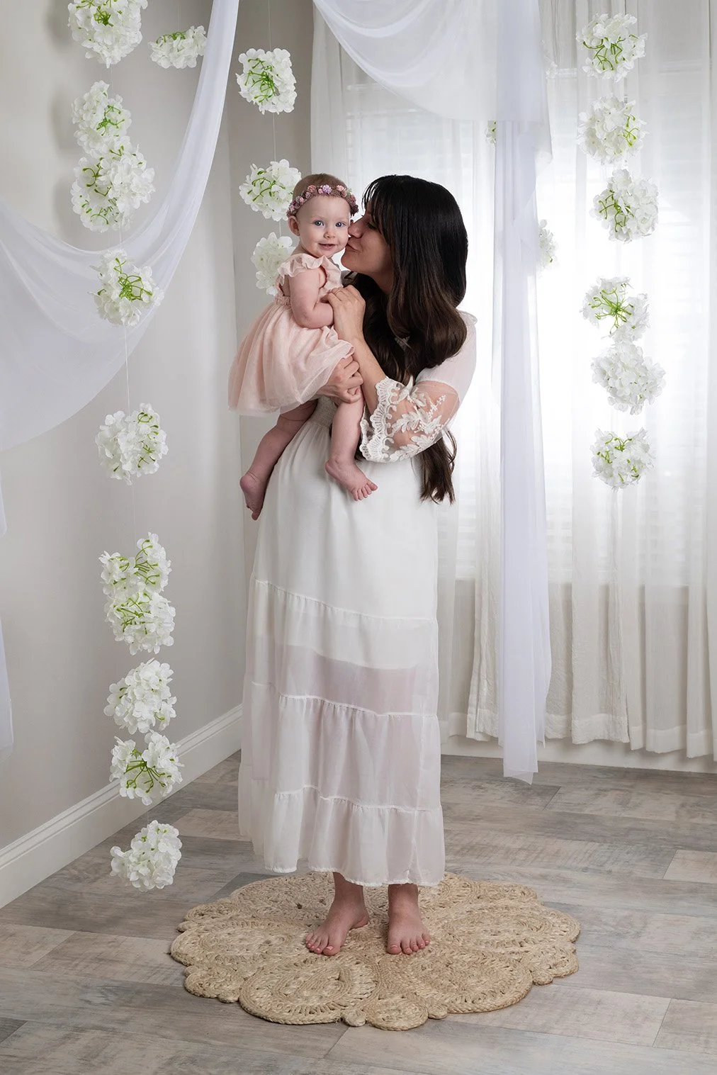 A woman holding a smiling baby girl in a room decorated with white curtains and hanging white flowers, standing on a beige braided rug.