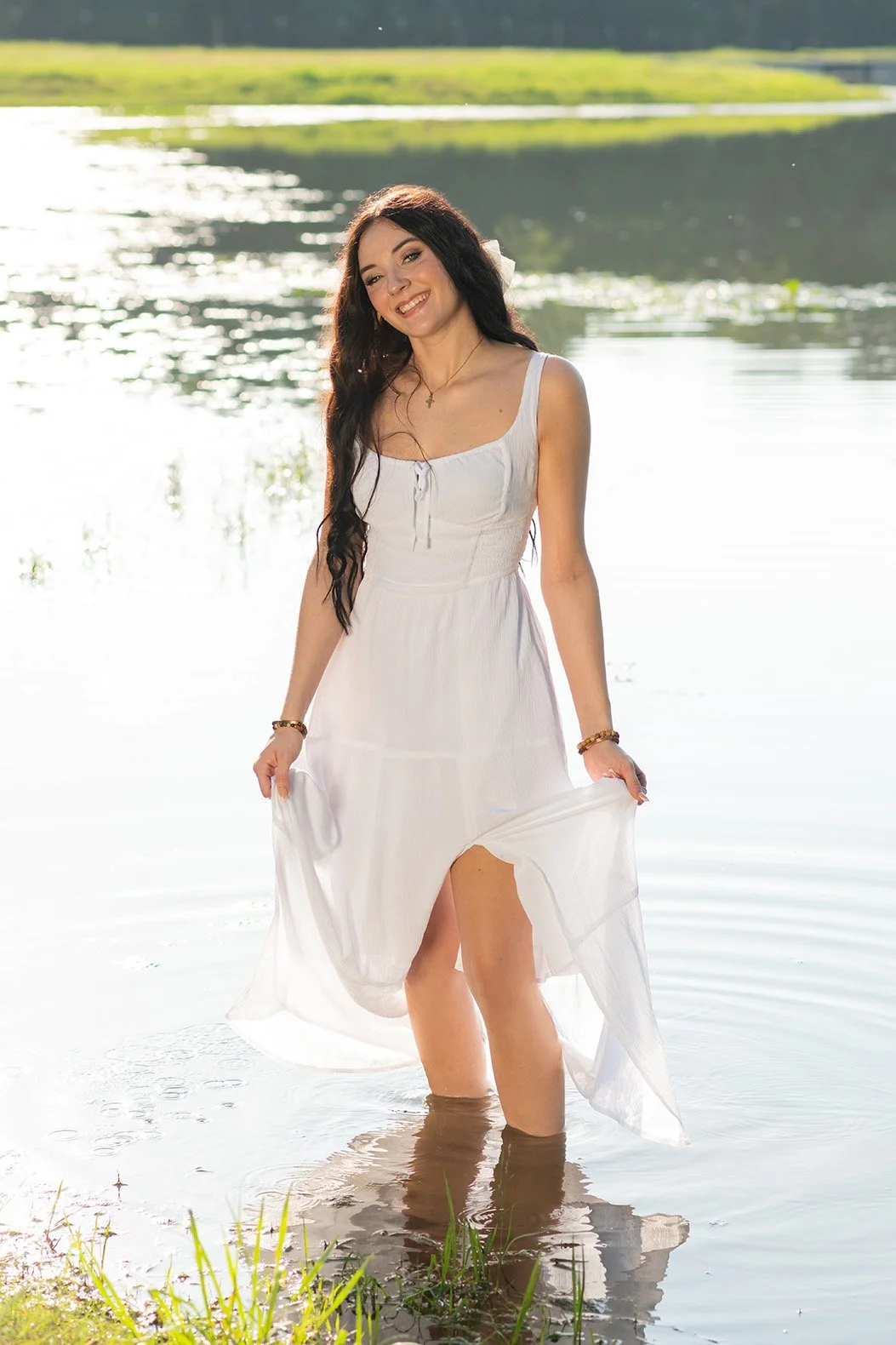 A woman in a white dress standing in shallow water near a lake, smiling and holding the hem of her dress.