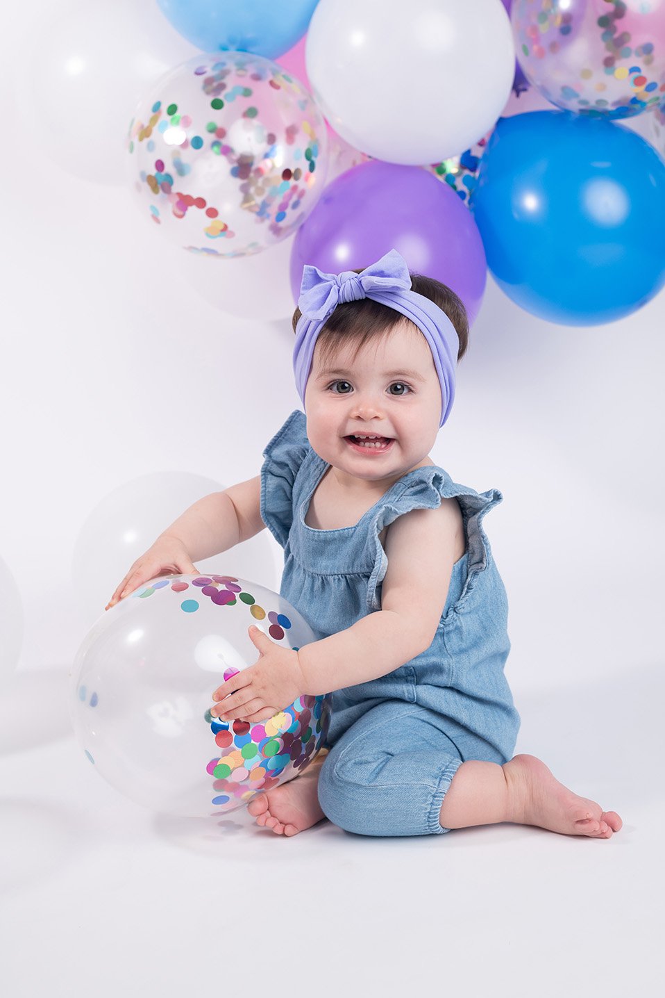 A smiling baby girl with a purple headband, wearing a denim romper, playing with a clear balloon filled with colorful confetti, surrounded by purple, blue, and clear balloons with confetti inside, against a white background.