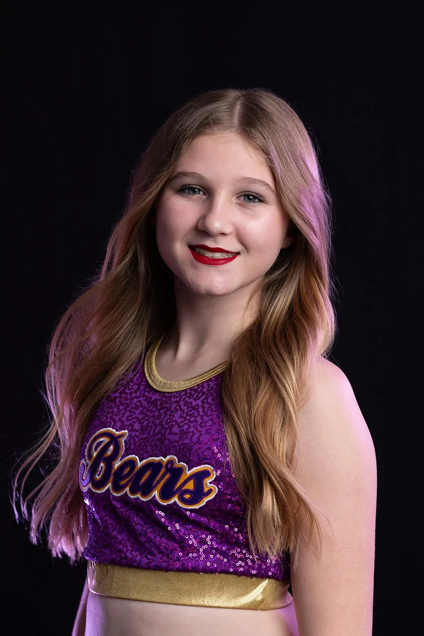 Young girl with long, wavy blonde hair wearing a purple cheerleading top with gold accents and the word 'Beavs' written on it, smiling against a black background.