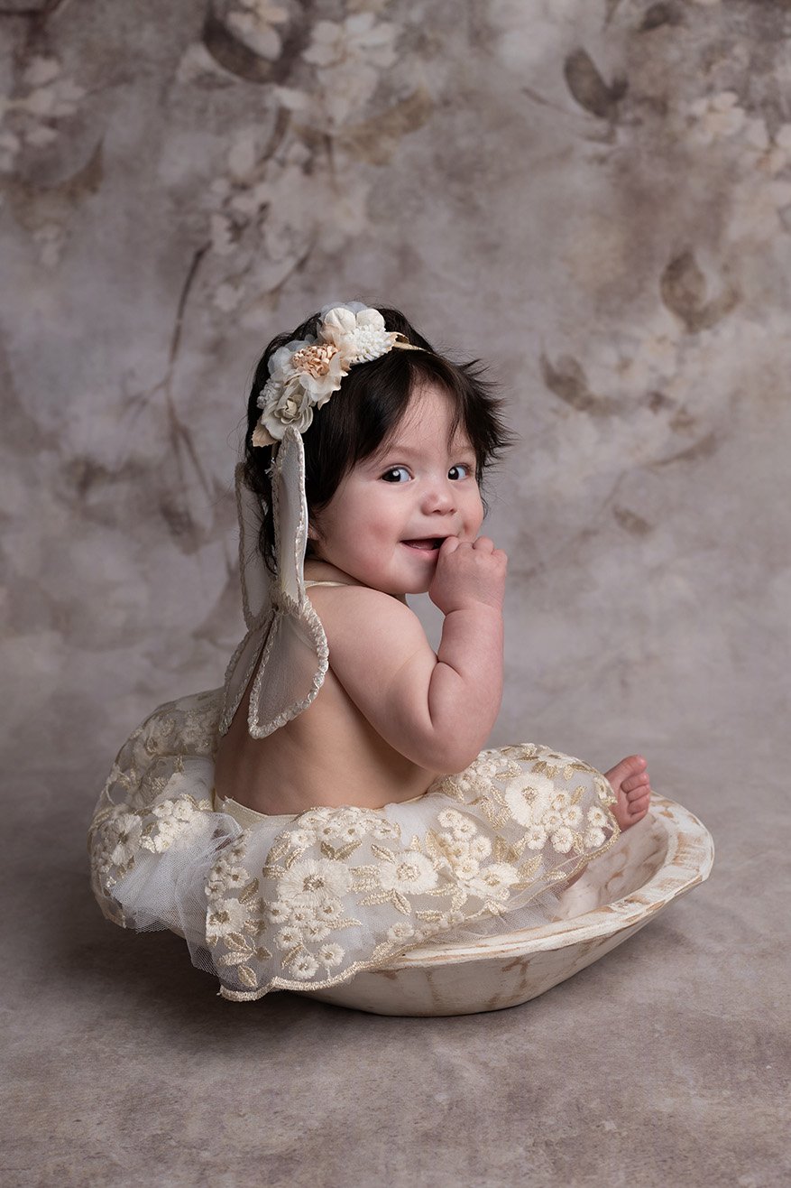 A baby with dark hair, dressed in cream-colored floral lace clothing with a headband, sitting in a shallow bowl against a neutral textured background.