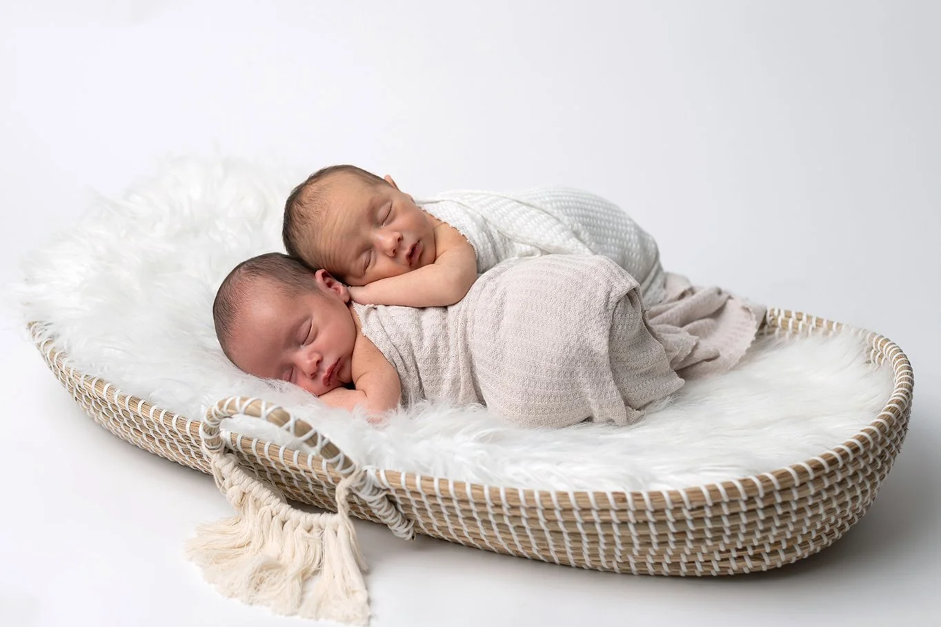 Two sleeping newborn babies lying on their sides on a soft, fluffy white blanket inside a woven basket.