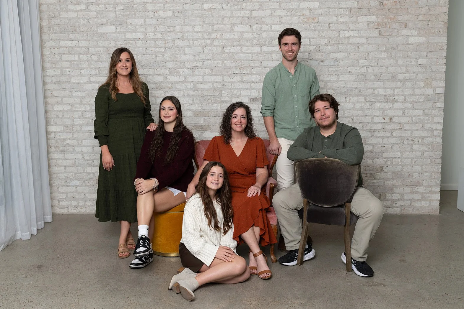 Group of seven people, including women and men, gathered in a room with a white brick wall background, posing for a portrait.