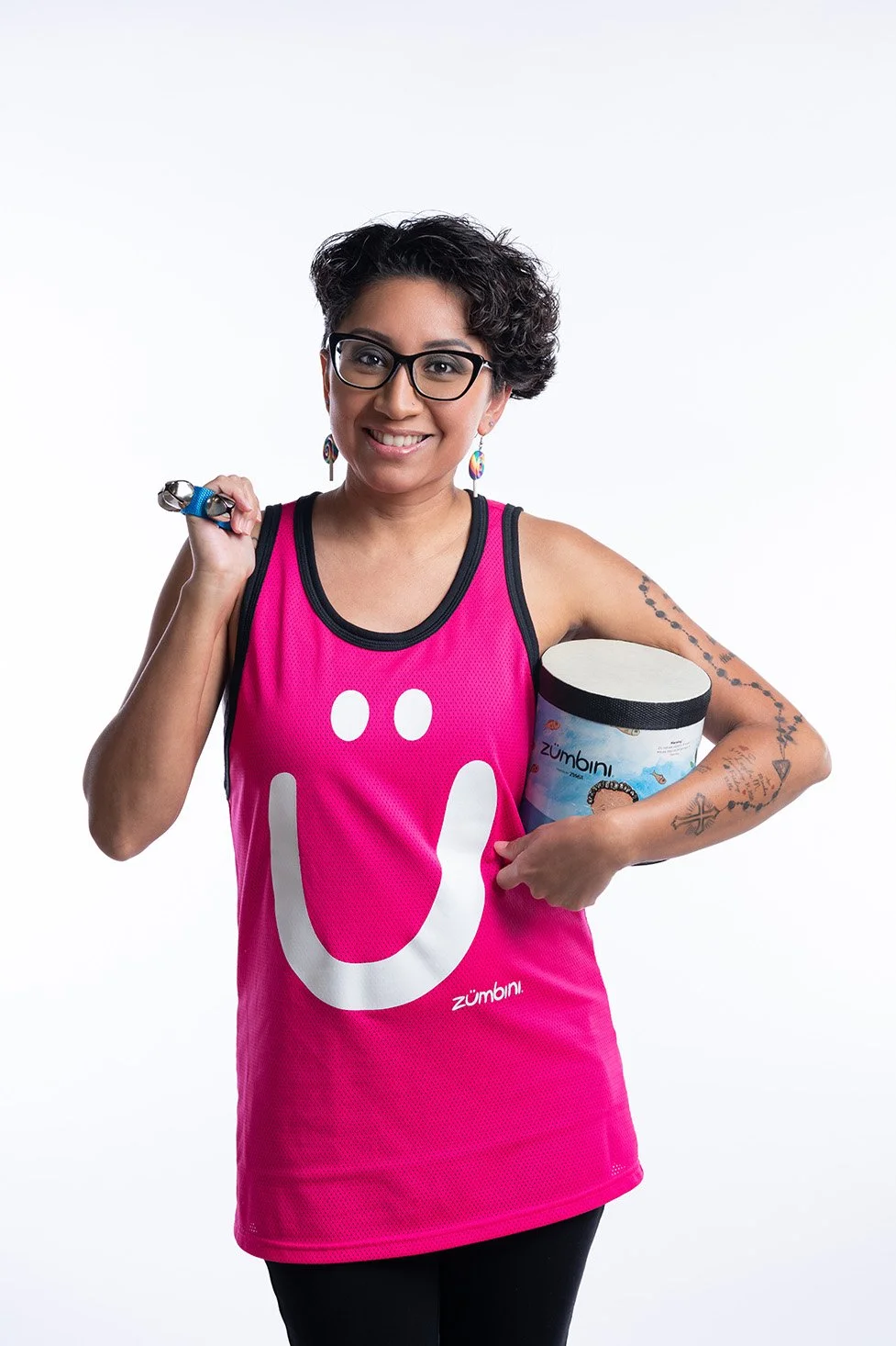 A woman with short curly hair, wearing glasses and a pink fitness dress with a smiley face design, holds a container of Zümbini ice cream in one hand and a small dumbbell in the other.