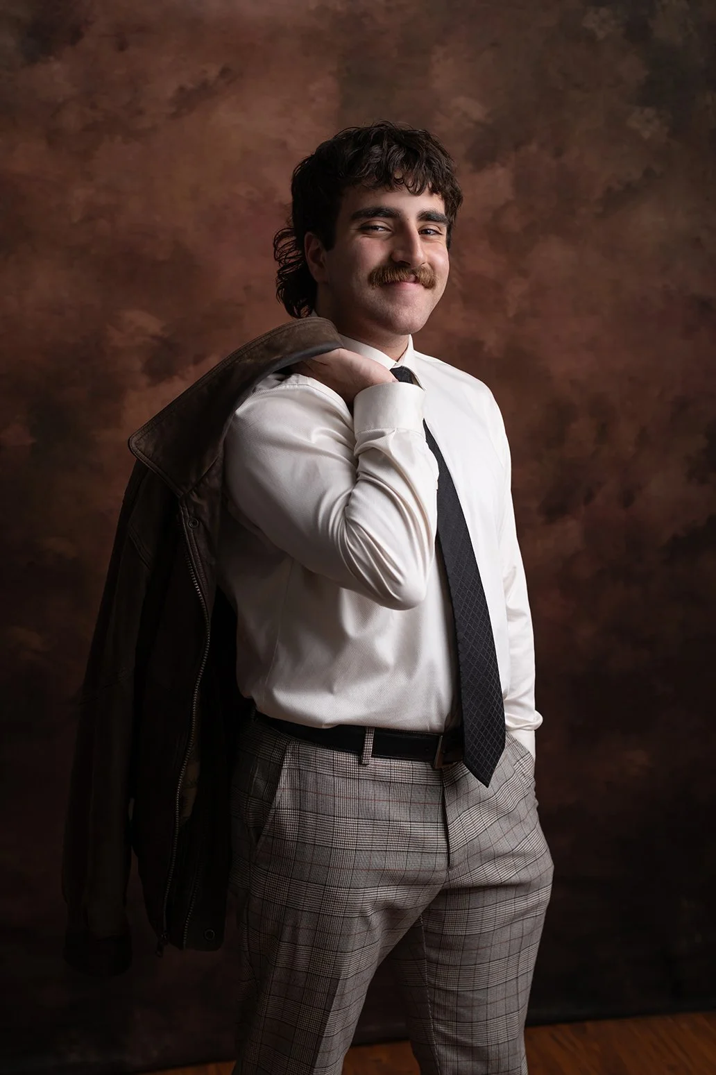 A man in vintage attire with a mustache, white shirt, black tie, plaid pants, holding a leather jacket over his shoulder and smiling.