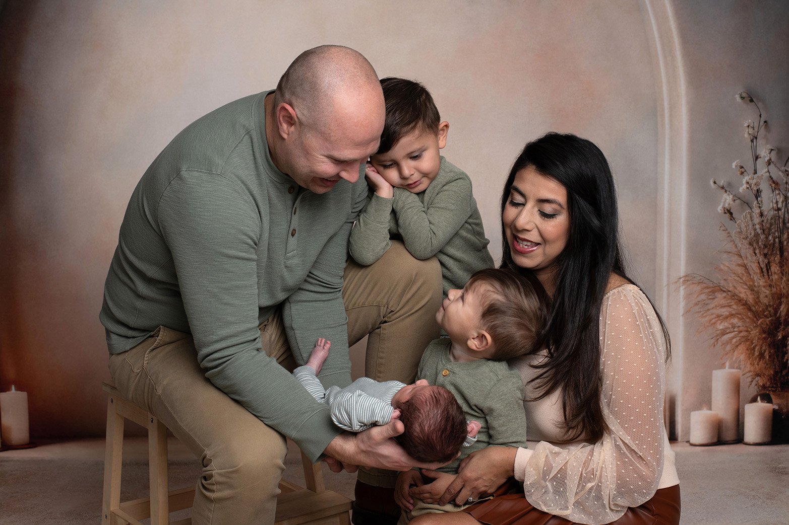 Family of five with two young children, a mother, father, and another child, sitting together indoors, looking at and holding a newborn baby.