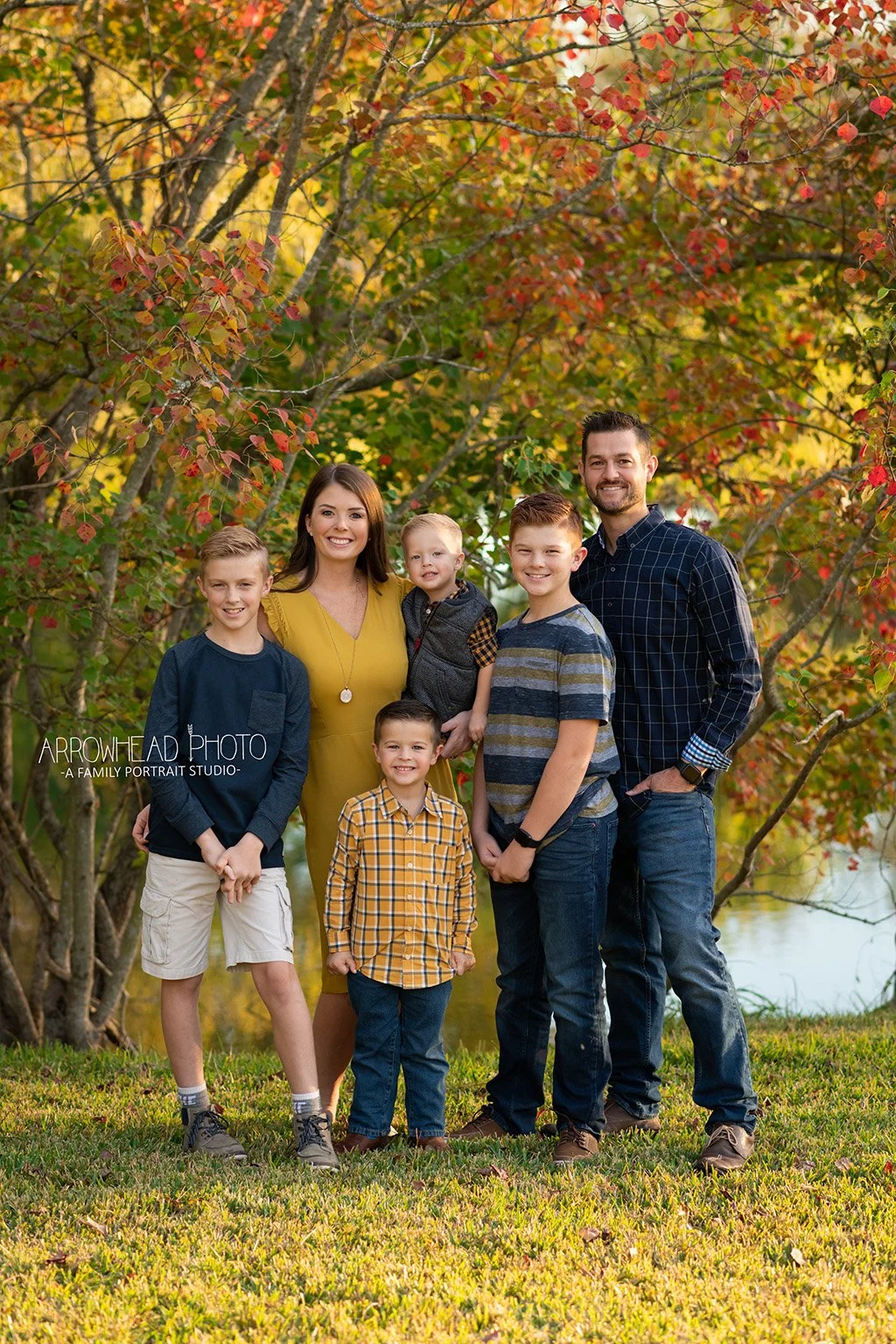 Family of six posing outdoors during fall, surrounded by colorful autumn trees with red, orange, and yellow leaves, standing on green grass near water.