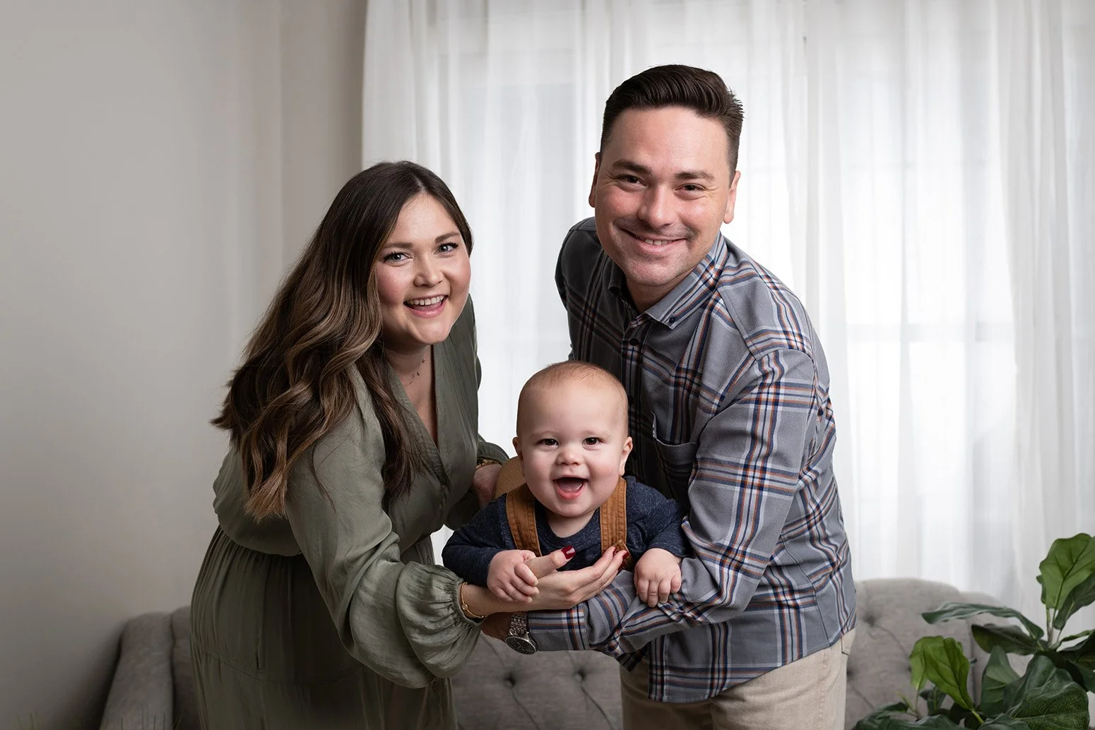 Happy family of three with a woman, man, and baby smiling indoors near a window with white curtains and greenery.