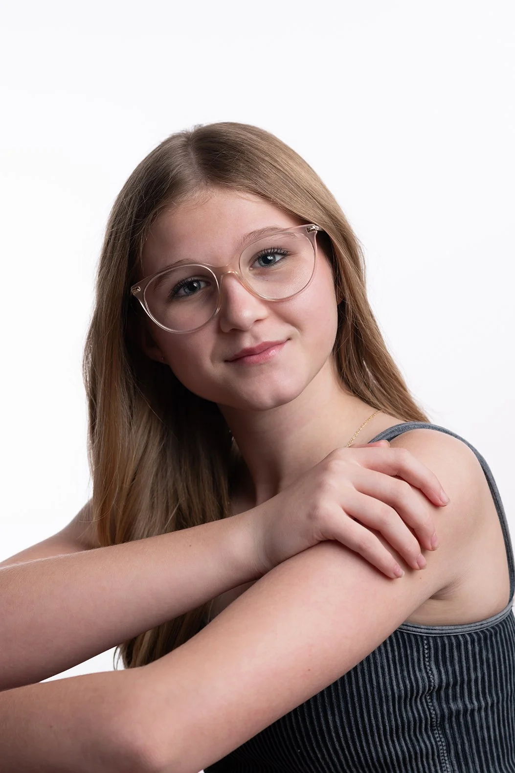 A young girl with long red hair wearing glasses and a gray tank top, gently touching her shoulder with a calm expression against a plain white background.
