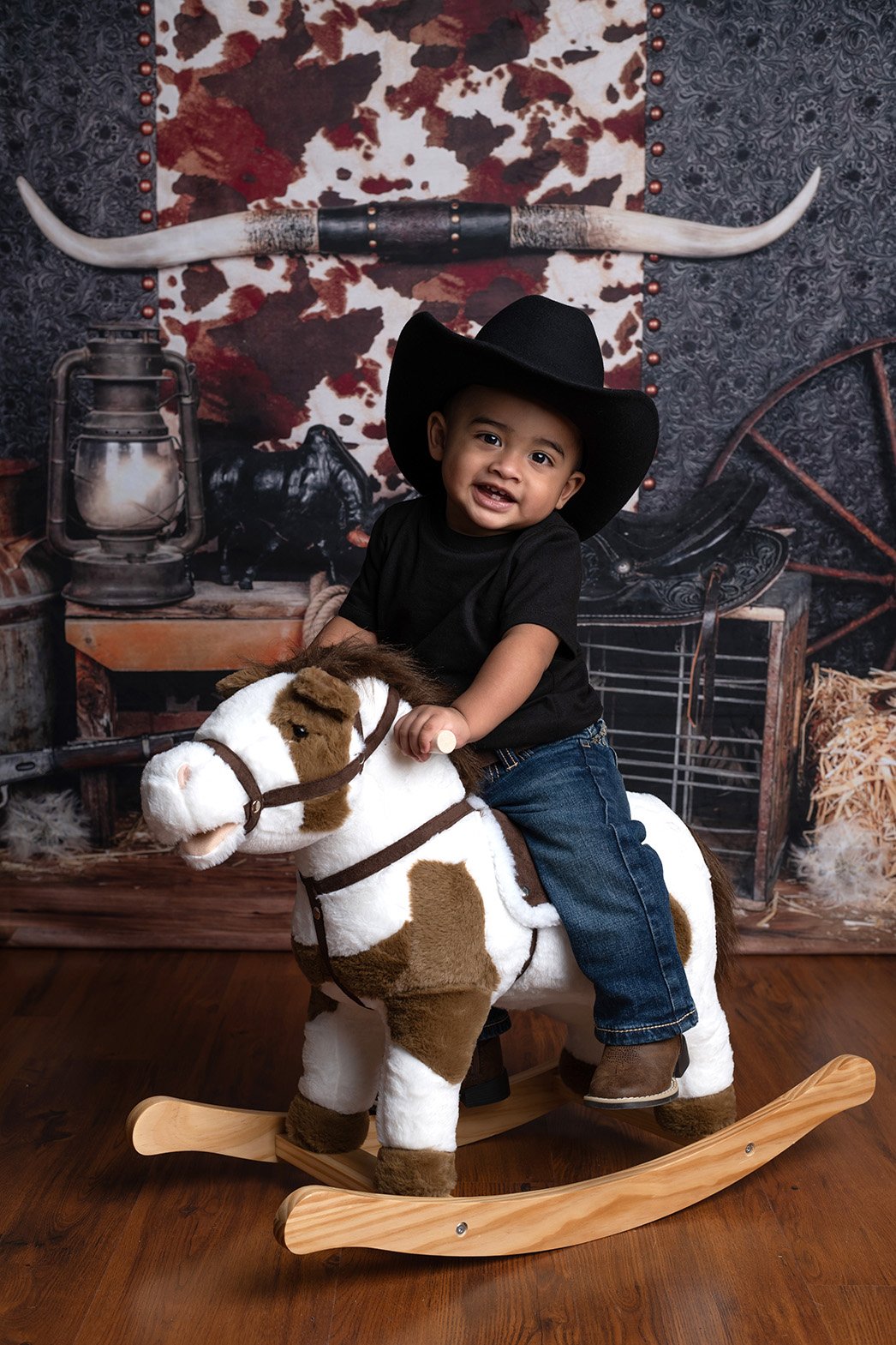 A young boy wearing a black cowboy hat and black shirt riding a plush rocking horse in a Western-themed setting.