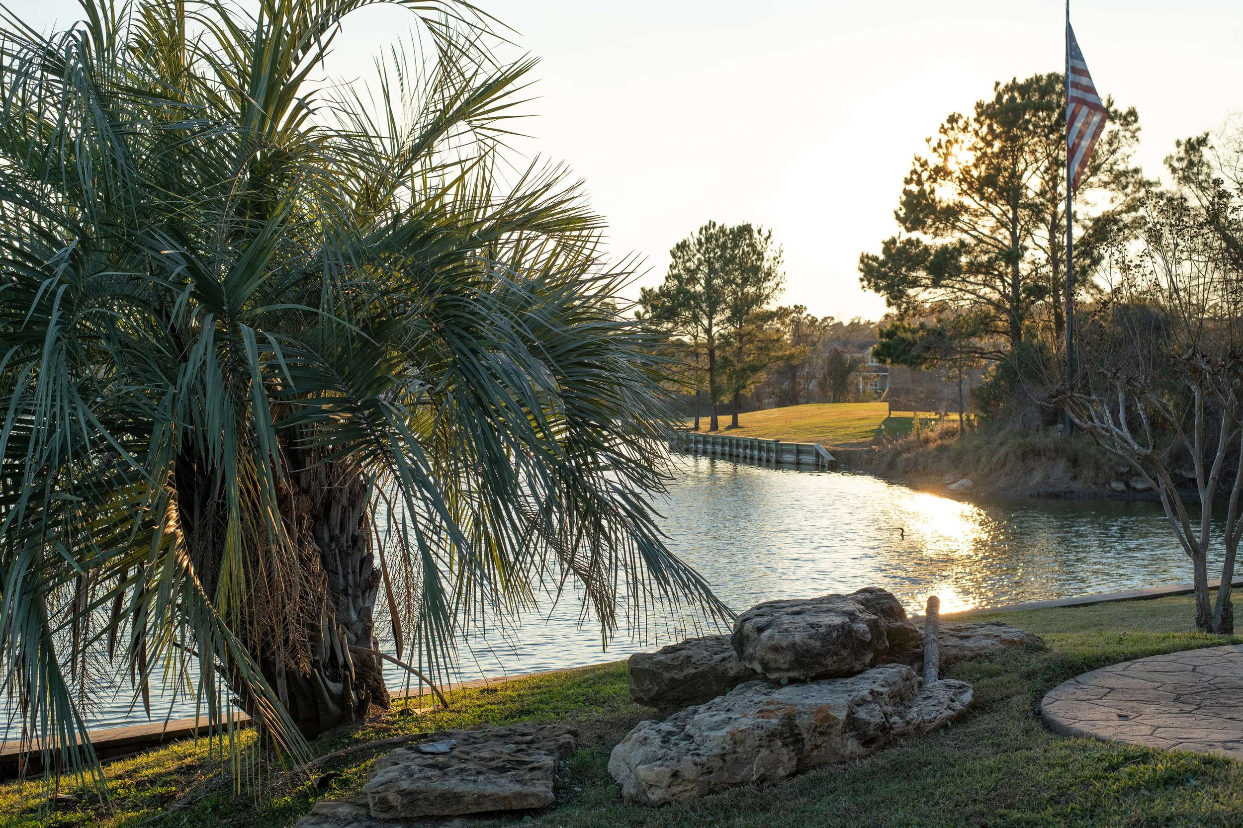 Sunset over a river with trees, rocks, grass, and an American flag.