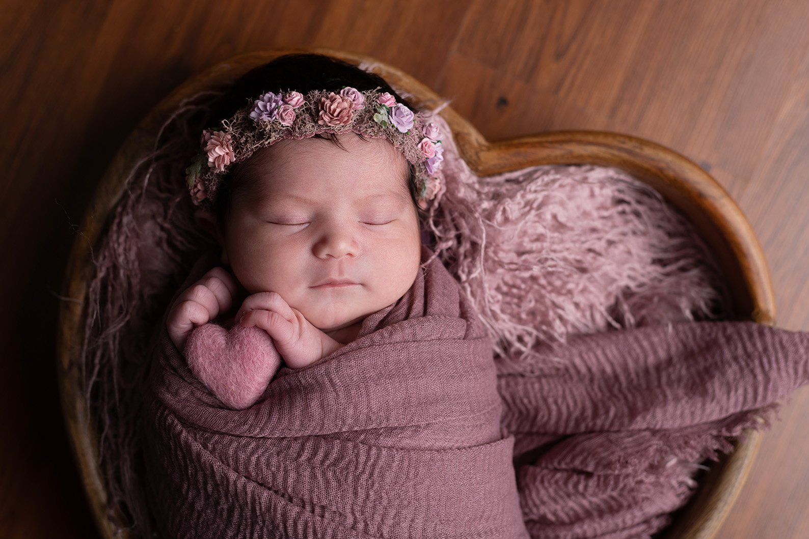 A newborn baby girl sleeping peacefully, wearing a pink floral headband, wrapped in pink and brown blankets, lying in a wooden cradle.