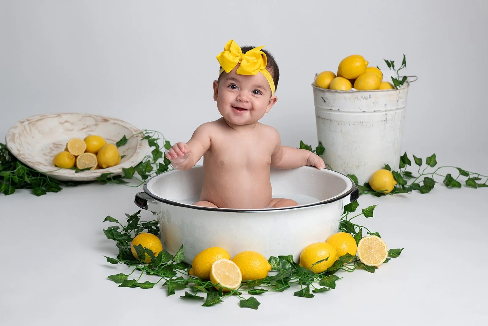 A baby sitting in a white basin filled with water, surrounded by lemons and green leaves, with more lemons in a white bucket and on a white plate in the background.