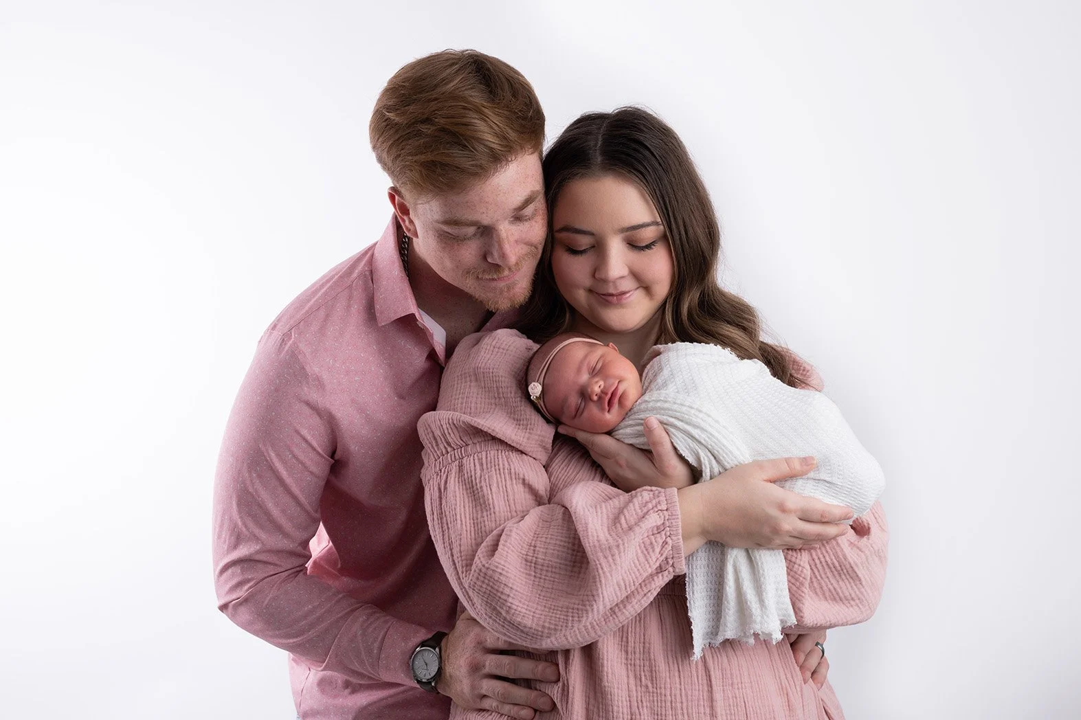 A family of three holding their newborn baby, smiling gently, with a plain white background.