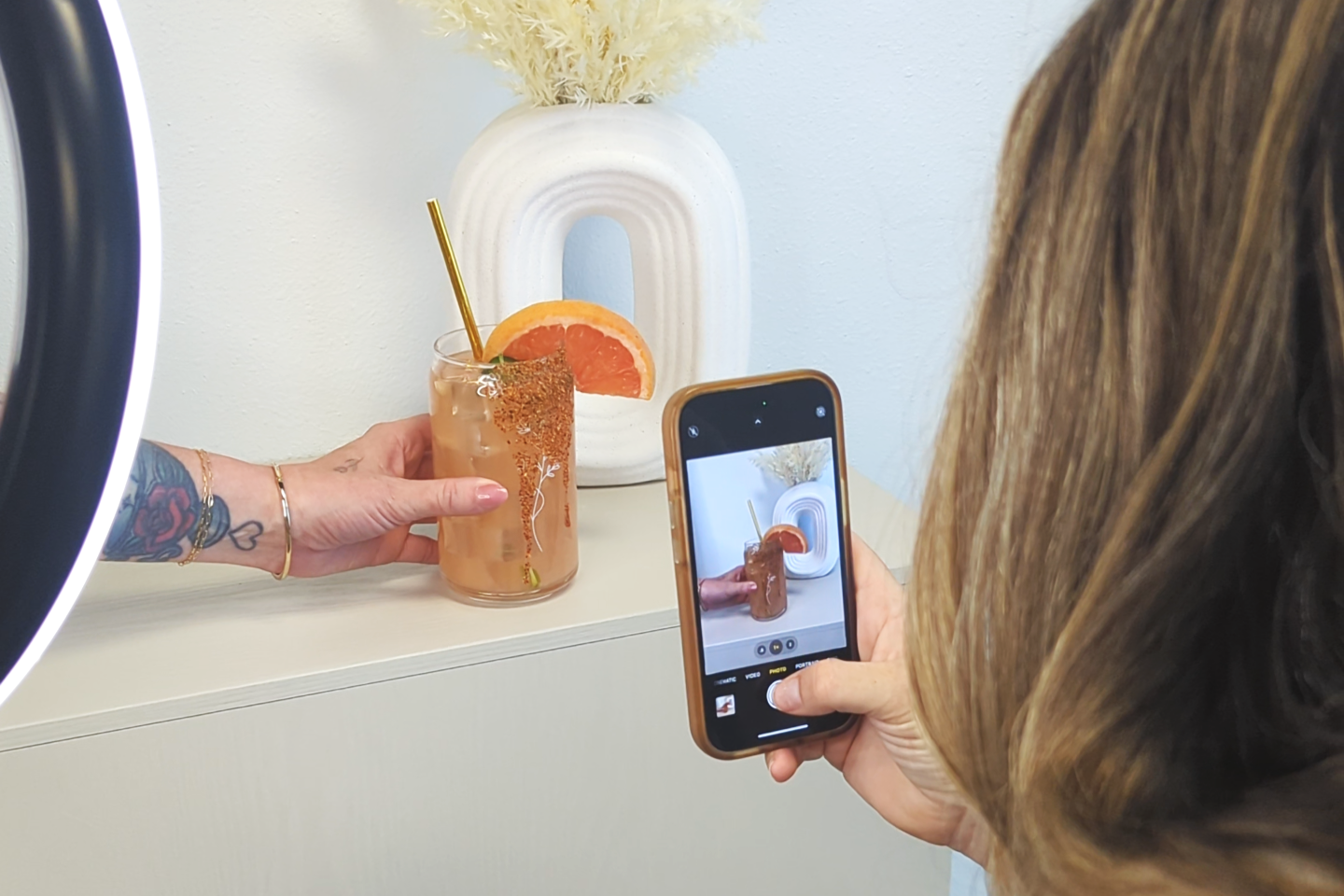 A person with long brown hair is taking a photo of a grapefruit cocktail with a smartphone. The cocktail is garnished with a grapefruit slice and a straw, and is in a glass decorated with a white tree design. In the background, there is a white abstract vase with dried flowers.