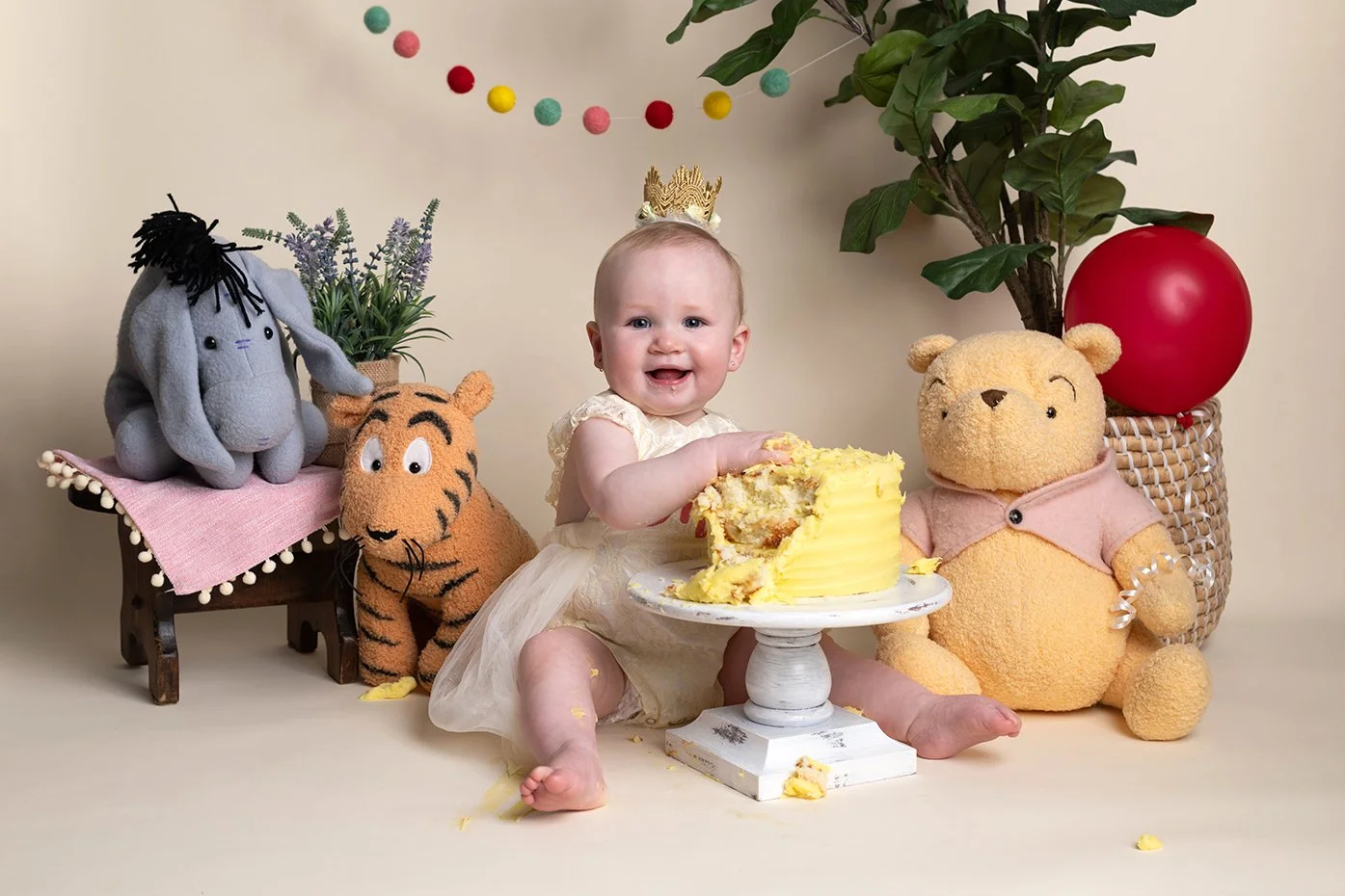 A baby girl sitting on the floor with a partially eaten yellow birthday cake on a white cake stand. She wears a cream-colored dress and a small golden crown, smiling at the camera. Behind her are plush toys of Eeyore, Tigger, and Winnie the Pooh, and
