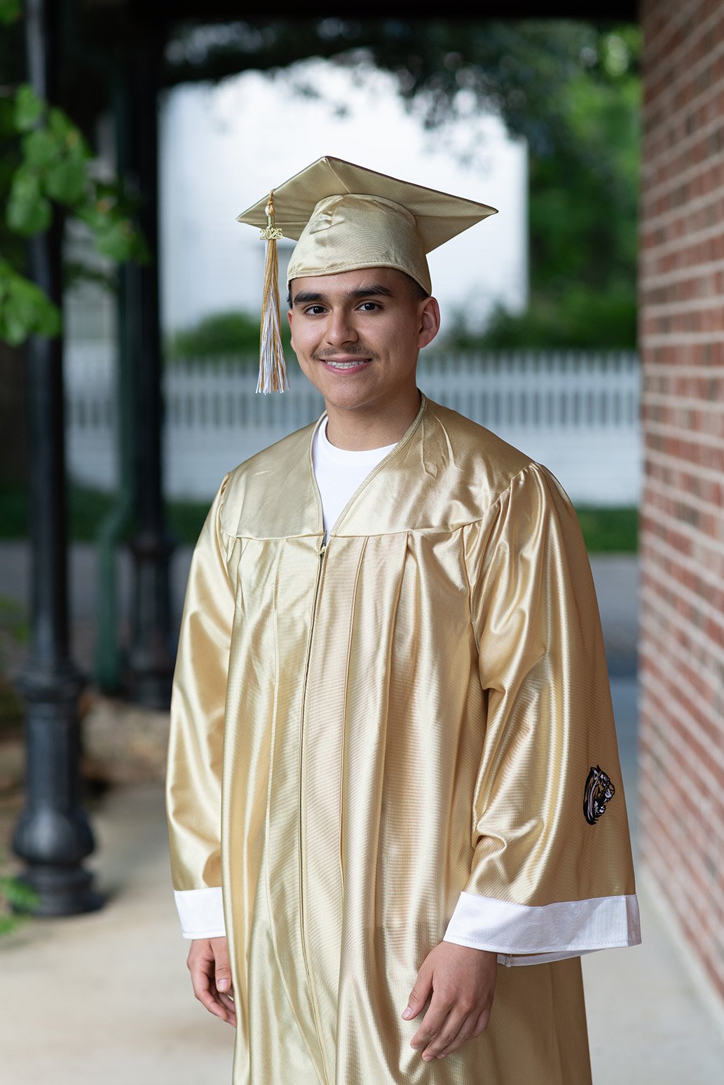 Young man in gold graduation gown and cap smiling outdoors with a brick wall and white fence in the background.