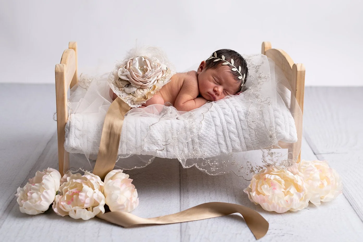 A newborn baby girl is sleeping on a small white bed, dressed with a lace blanket and adorned with a headband and a large flower on her back, surrounded by flowers and ribbons