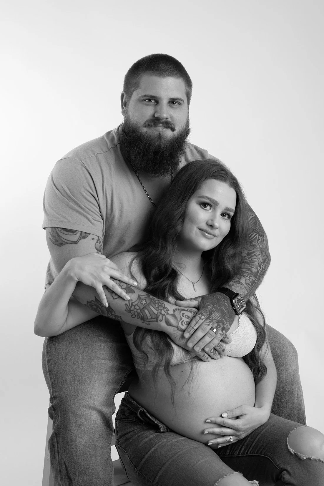 A pregnant woman with long wavy hair and tattoos on her arms and stomach, sitting on a stool, with a bearded man standing behind her, embracing her, both smiling, in a studio setting.