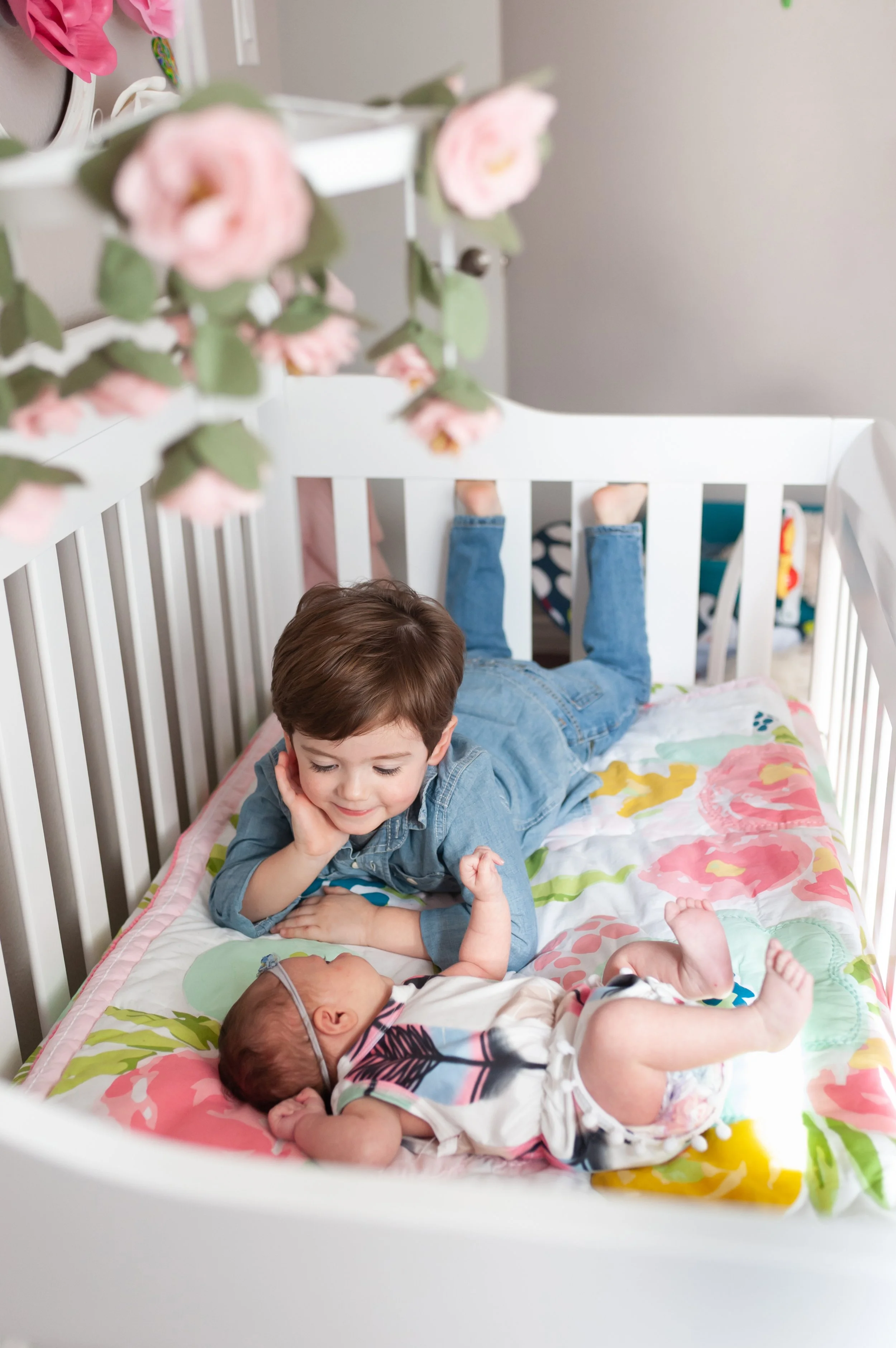 A young boy lying on his stomach in a crib, smiling and looking at a newborn baby lying on the same bed.