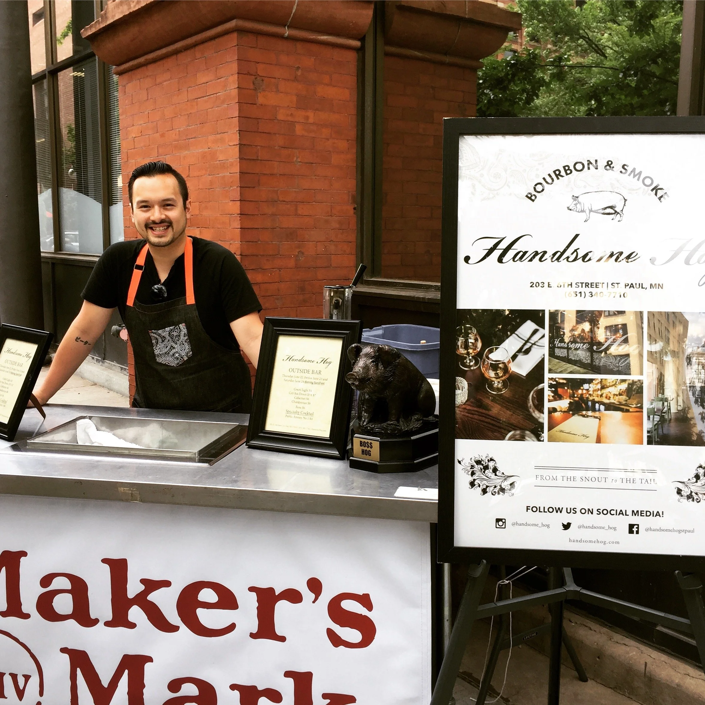 A man smiling at a food stand with a black apron and orange straps, next to a black pig statue and a sign for Handsome Hog at an outdoor market or festival.