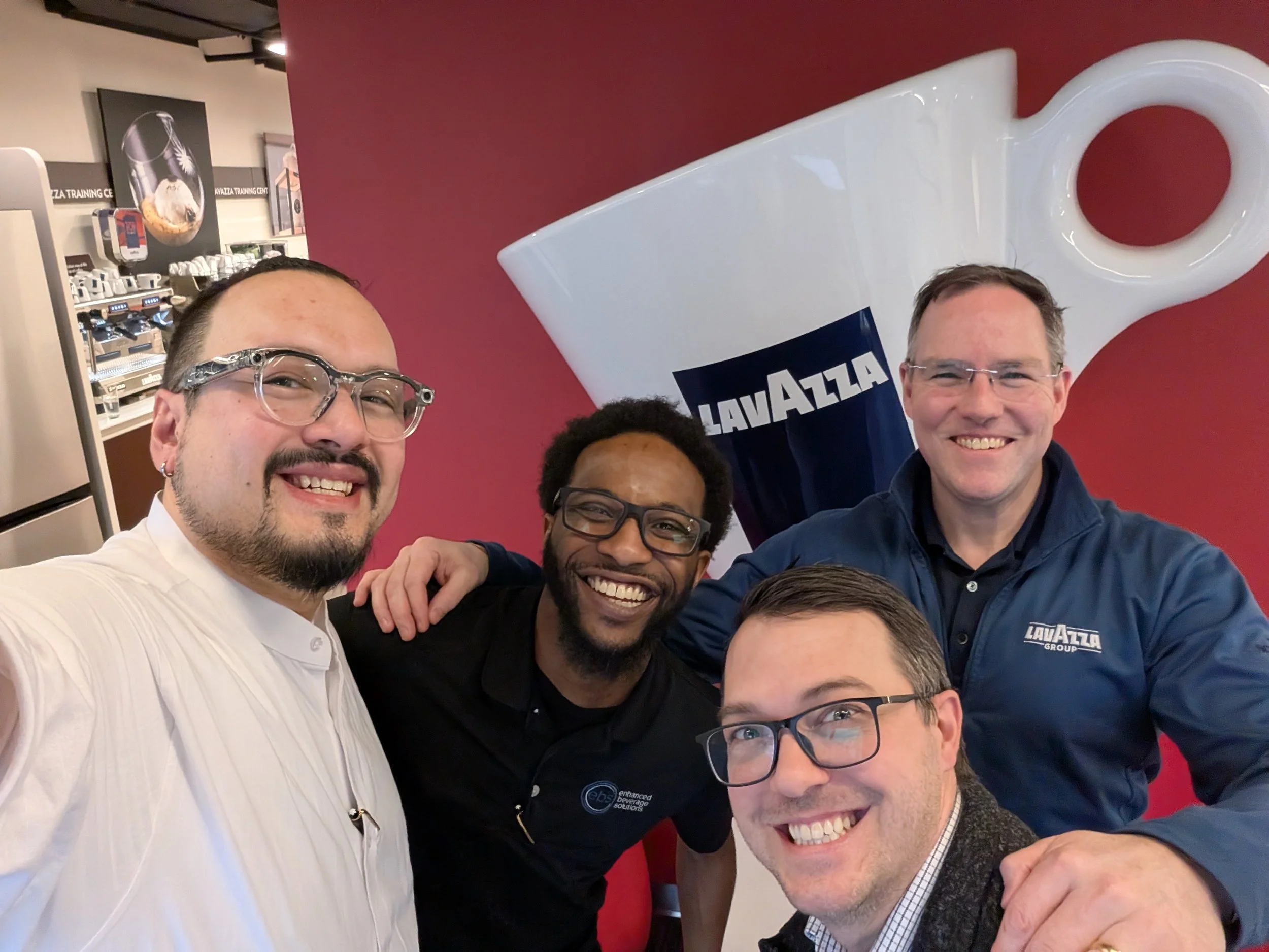 Four smiling men taking a selfie in front of a large coffee mug with the Lavazza logo, standing in a coffee shop or cafe environment.