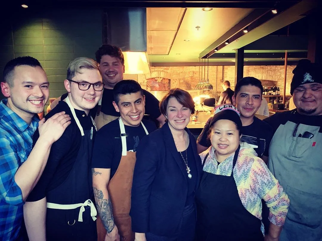 Group of people smiling and posing together in a restaurant or cafe, some wearing aprons, indicating they might be staff or chefs.