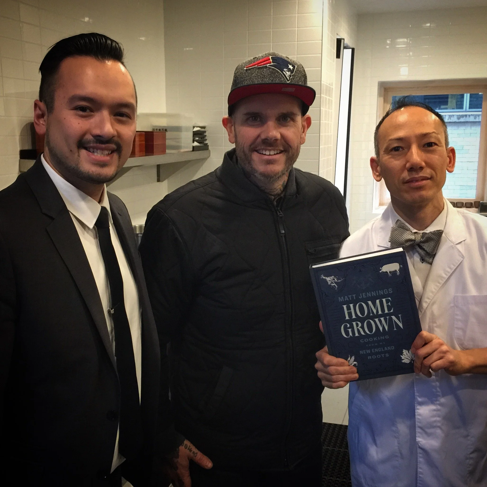 Three men standing together indoors, with one holding a book titled 'Home Grown' by Matt Jennings, in a white-tiled room with a window and shelves in the background.