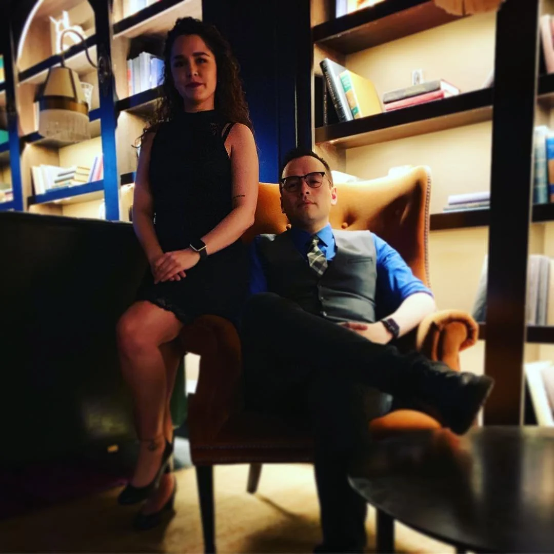 A woman in a black dress stands next to a seated man in a blue shirt, gray vest, and plaid tie, both in a room with wooden bookshelves filled with books, some standing and some lying flat, and some decorative objects. The woman has curly hair and a t