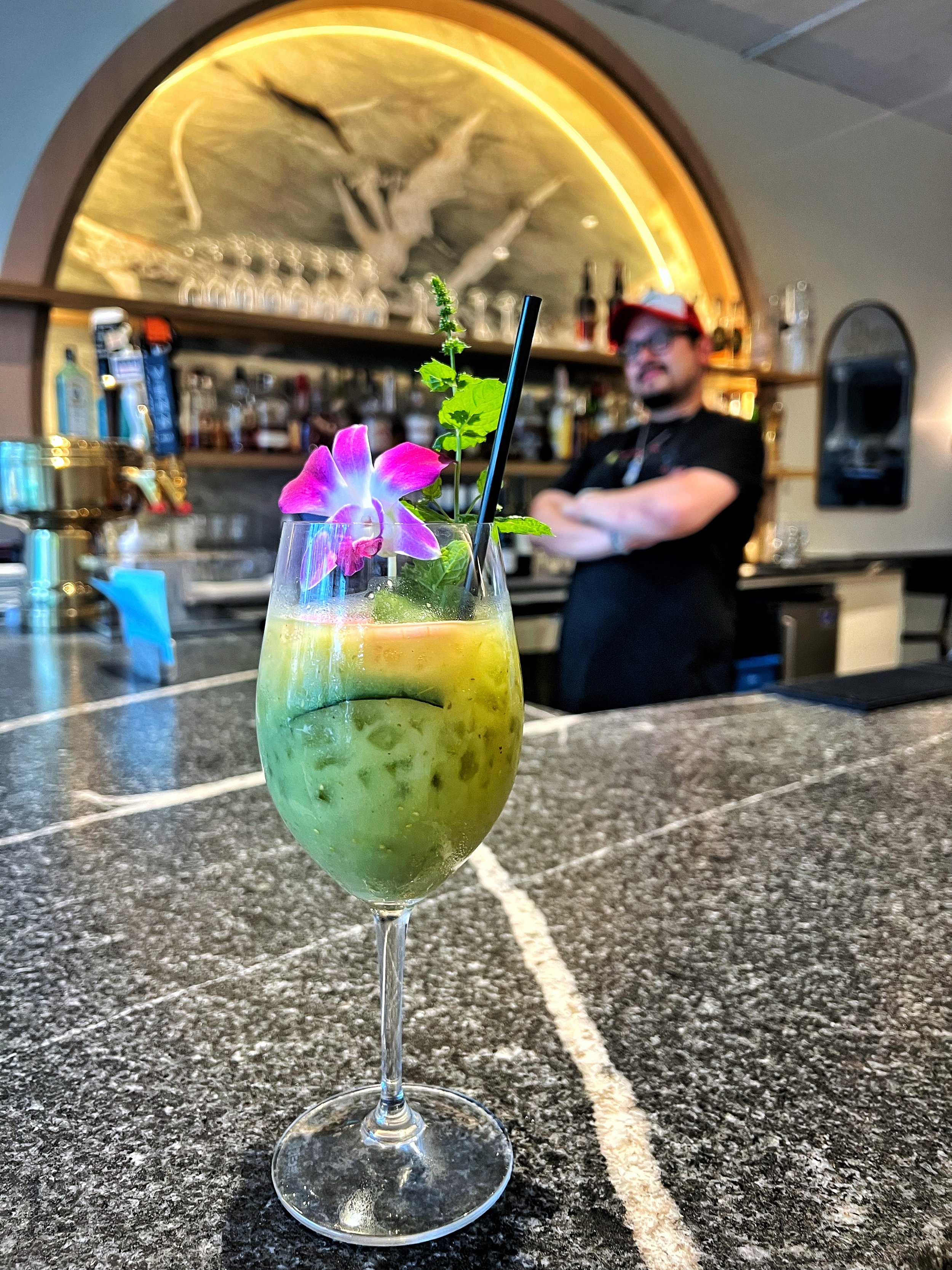 A tropical cocktail garnished with a purple flower and mint leaves, served in a tall glass with a black straw, placed on a textured bar counter.