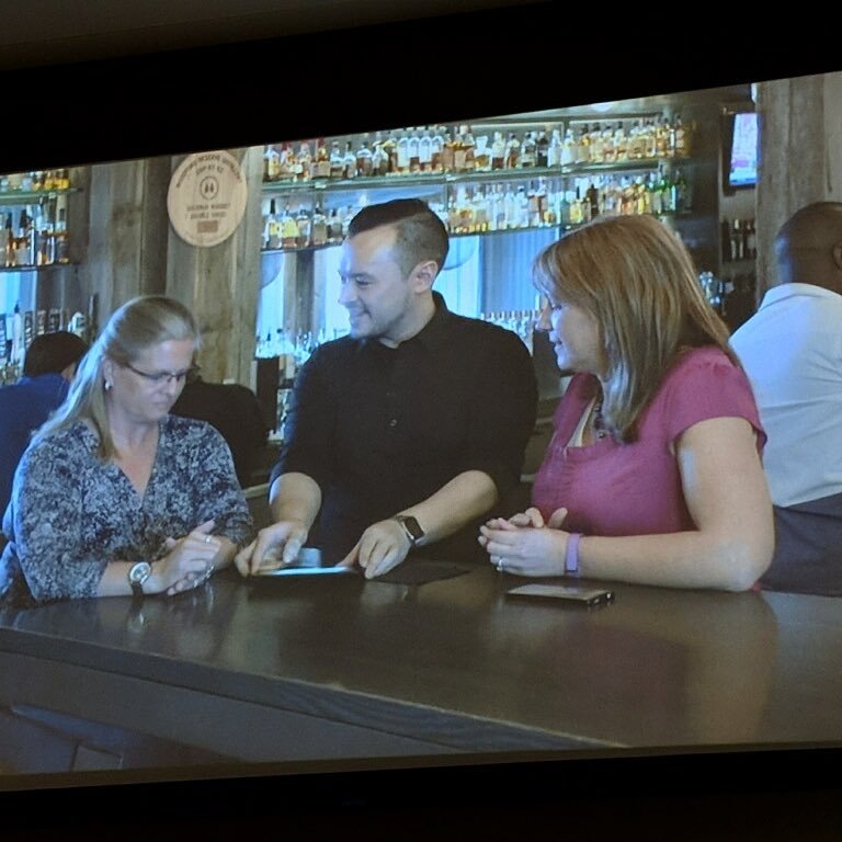 Three people are sitting at a bar, with a woman in glasses on the left looking at her phone, a man in the middle smiling at a phone, and a woman in a pink shirt on the right looking at the phone. There are bottles on shelves in the background.