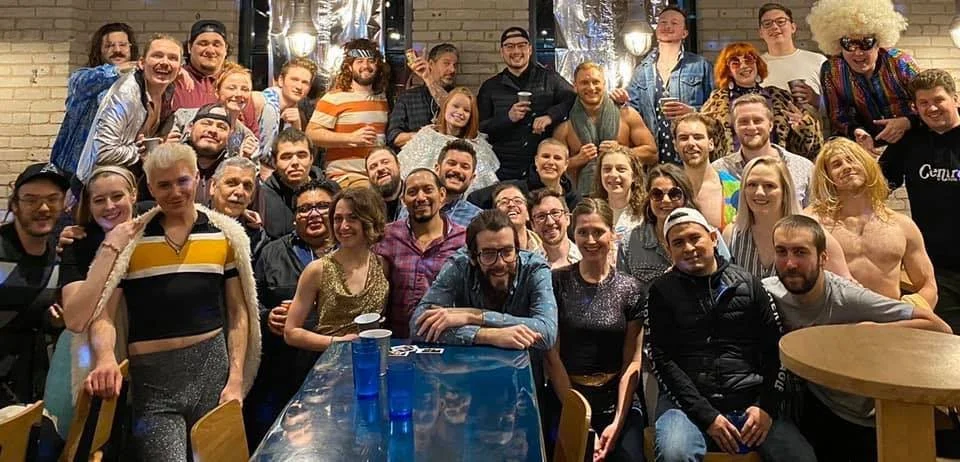 Group of people celebrating at a party, some wearing costumes and casual attire, posing together indoors with a brick wall and lighting in the background.