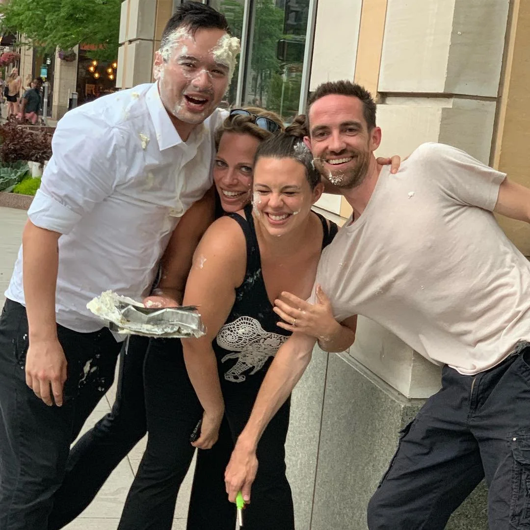 Group of four friends outside, celebrating with cake and cream on their faces, smiling and having fun.