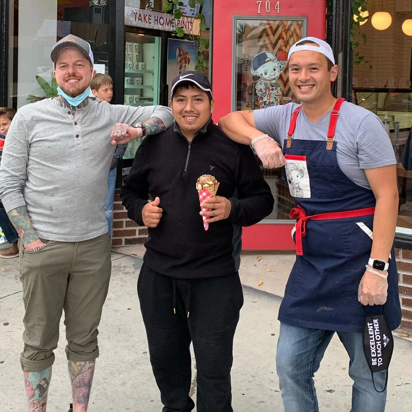 Three men standing outside a shop, smiling, with the man in the center holding an ice cream cone. The man on the left has tattoos and is wearing a gray shirt, a cap, and a face mask around his neck. The man on the right is wearing an apron, a gray T-