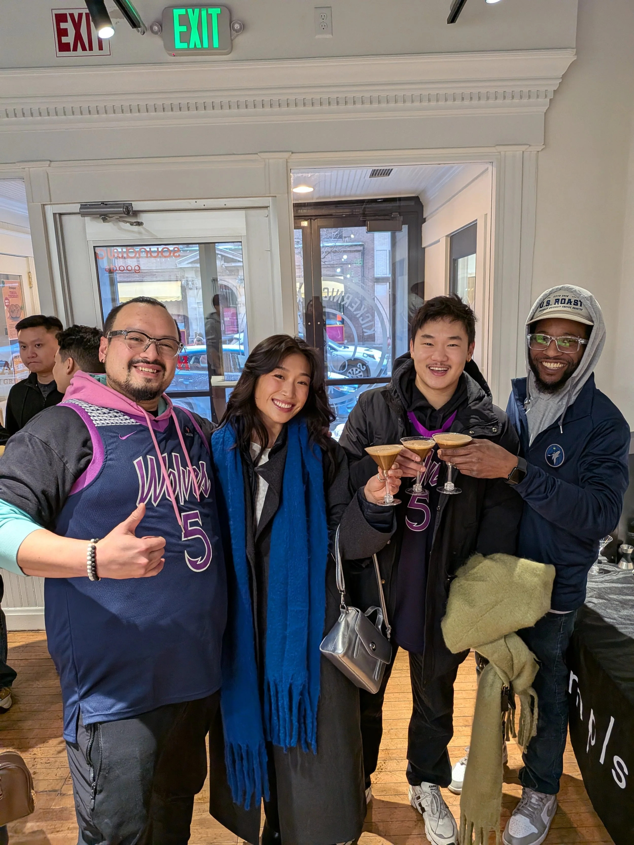Four people smile and hold up glasses of drinks at an indoor social event, with two men wearing Athletes in Action jerseys and a woman and man dressed casually. The background shows a glass door and window with a street view.