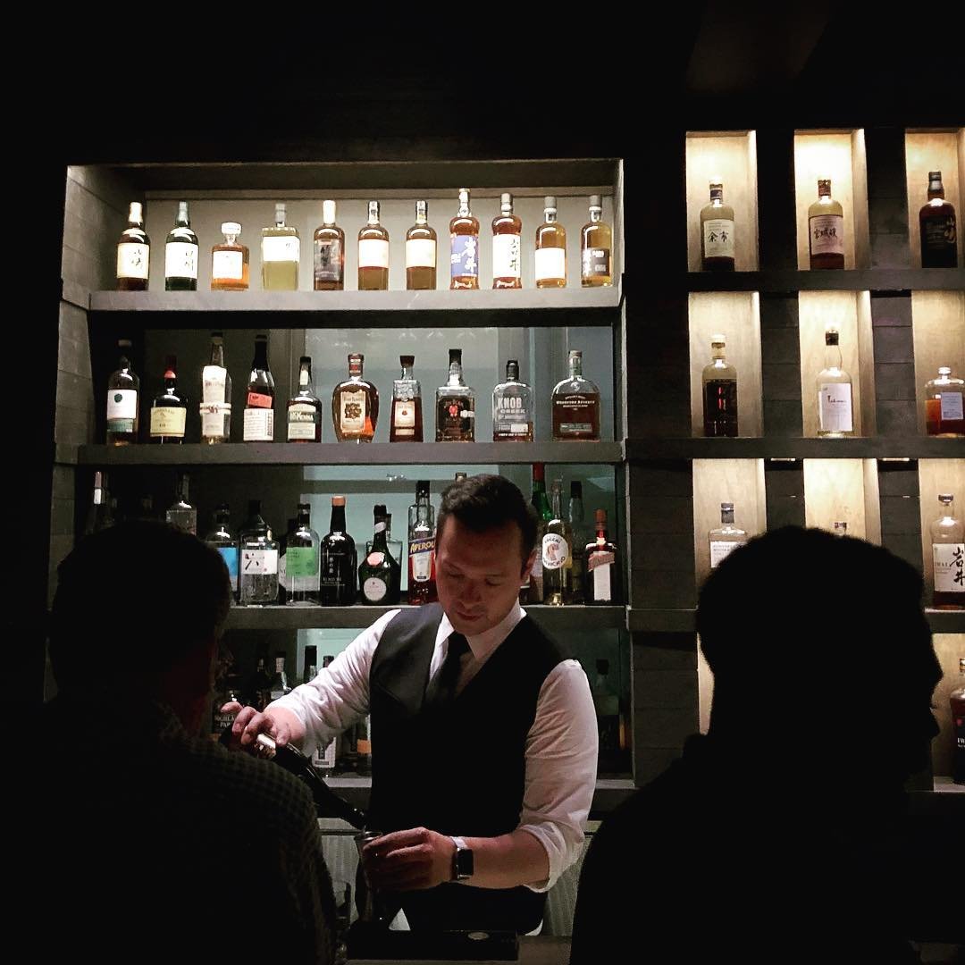 Bartender serving drinks to two customers at a bar with shelves of liquor bottles behind him.
