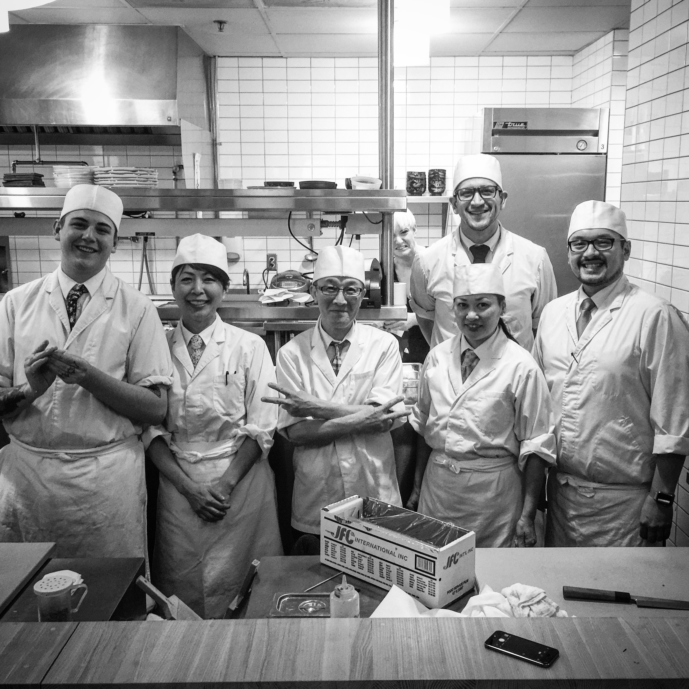 Group of seven chefs in a kitchen, all wearing white uniforms and hats, smiling for the camera.