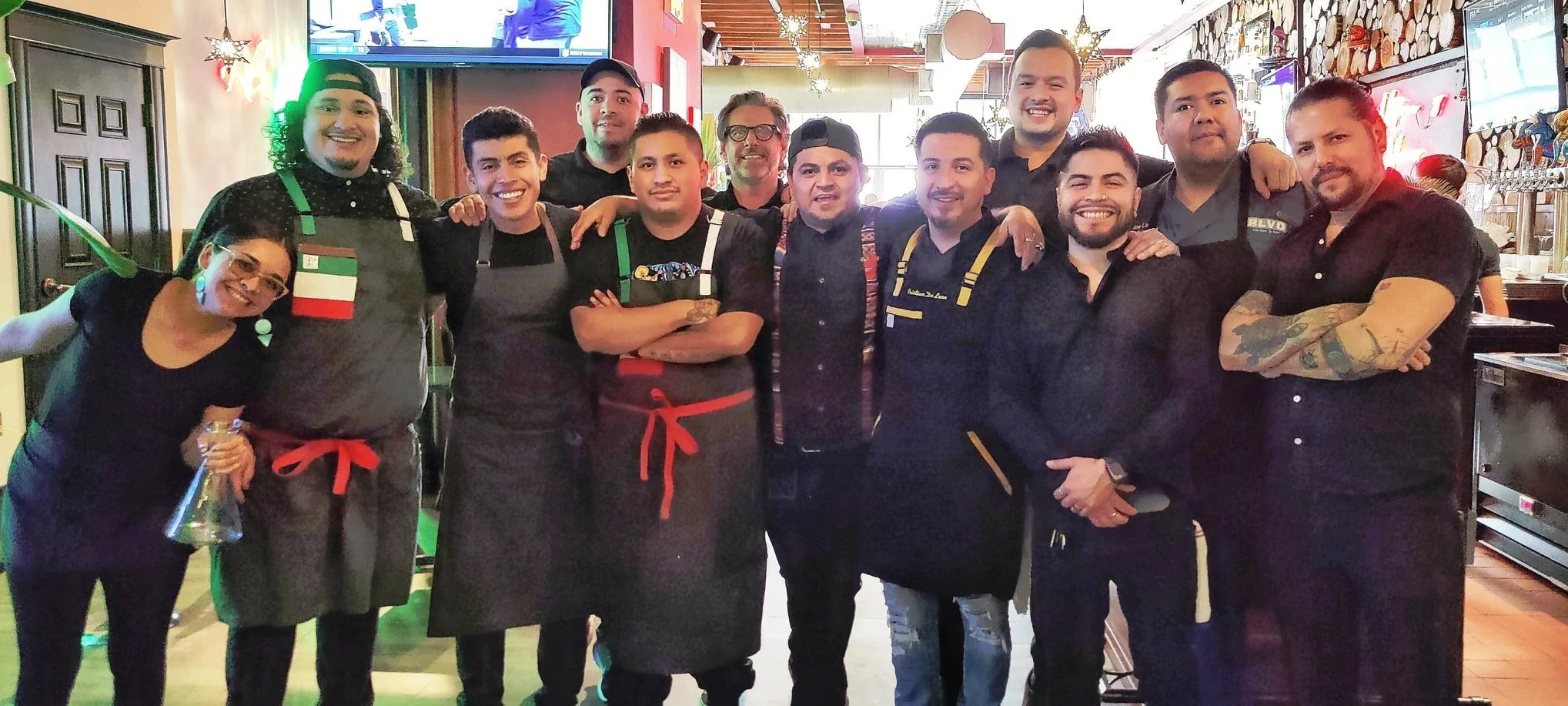 Group of twelve restaurant staff members posing together in a restaurant, some wearing aprons, some with tattoos, smiling, in front of a decorated dining area with wood accents and holiday decorations.