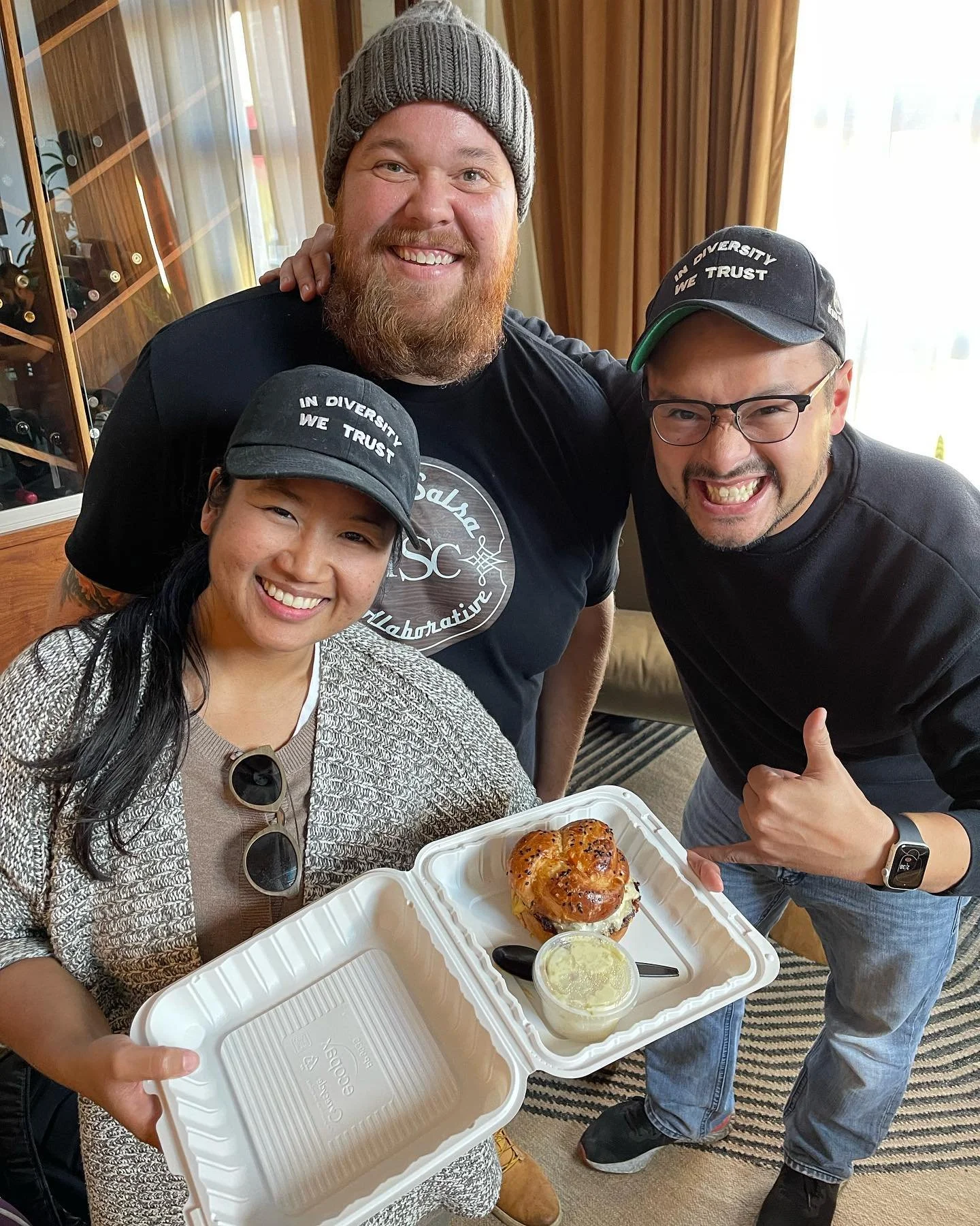 Three people smiling and celebrating, holding a takeout container with a baked item and a small cup of sauce or dip, inside a room with a wine rack and orange curtains.