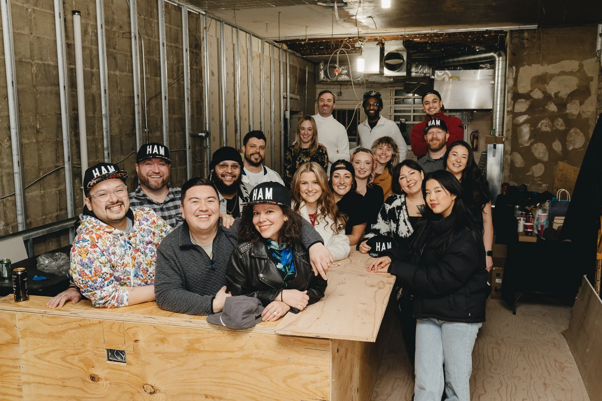 A diverse group of people gathered in a partially constructed room, smiling at the camera, some wearing hats with 'HAM' written on them.